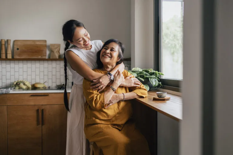 A Happy Beautiful Woman Hugging Her Mother While She Is Sitting In The Kitchen And Drinking Tea